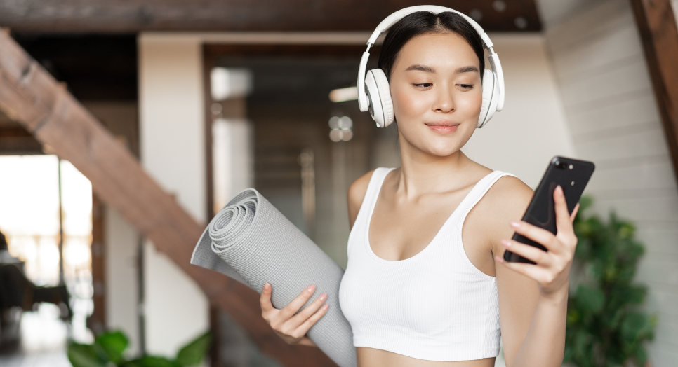 Woman with headphones looking at smart phone carrying yoga mat 